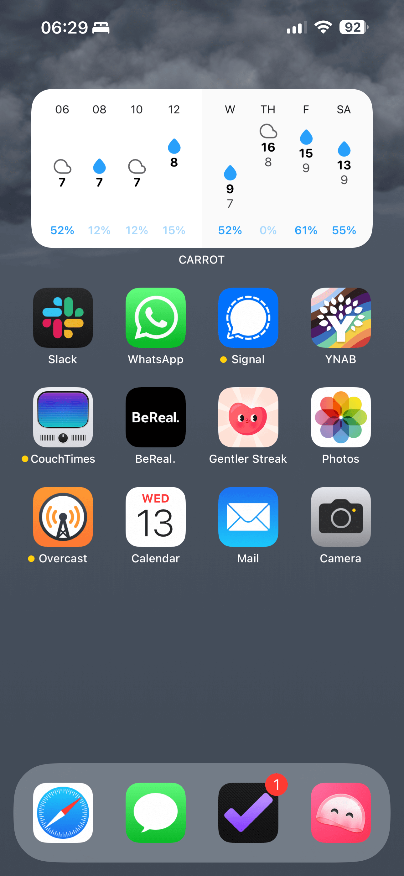 An iOS home screen with the weather background and a medium CARROT Weather widget at the top. After that comes three rows of apps: Slack, WhatsApp, Signal, YNAB, CouchTimes, BeReal, Gentler Streak, Photos, Overcast, Calendar, Mail, Camera. The Dock has four apps: Safari, Messages, OmniFocus and Manet.